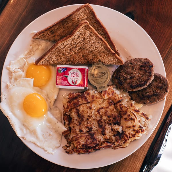 A soul food breakfast with two fried eggs, hash browns, sausage patties, and toast on a white plate.
