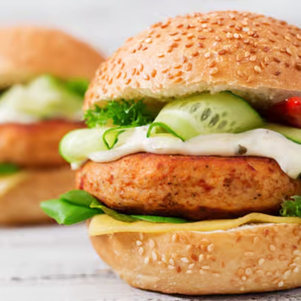 Two turkey burgers on sesame seed buns with cheese, lettuce, cucumber, and white sauce.